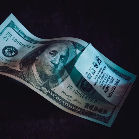 Close-up of a U.S. one hundred dollar bill with a fabric tag displaying care instructions. The bill is prominently featured and slightly curved.