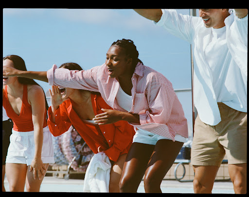 Group of young adults laugh outdoors on a sunny day. Women wear red and white tops and white shorts, man wears a white shirt and tan shorts.