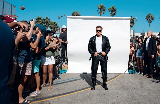 Arnold Schwarzenegger standing outdoors wearing black leather jacket, surrounded by people taking pictures on sunny day.