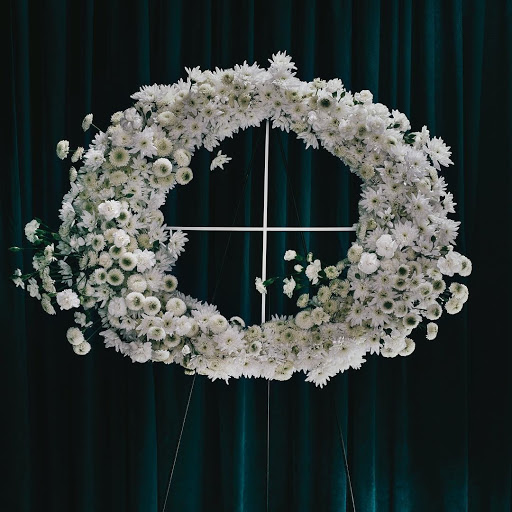 Floral wreath crafted from white daisies and supported by a white metal stand against a dark teal draped fabric backdrop.