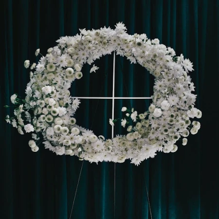 Floral wreath crafted from white daisies and supported by a white metal stand against a dark teal draped fabric backdrop.