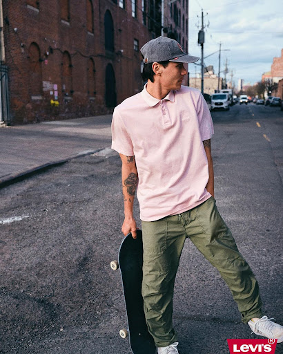 Man in pink polo and green pants holds skateboard on urban street near brick building, posing for fashion advertisement featuring Levi's logo.
