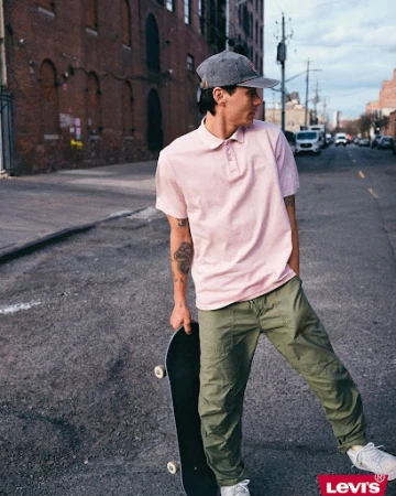Man in pink polo and green pants holds skateboard on urban street near brick building, posing for fashion advertisement featuring Levi's logo.