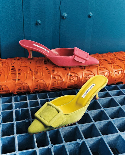 Two Manolo Blahnik mules in yellow and pink with signature buckle detail sit on a grid of orange and blue, showcasing high fashion.