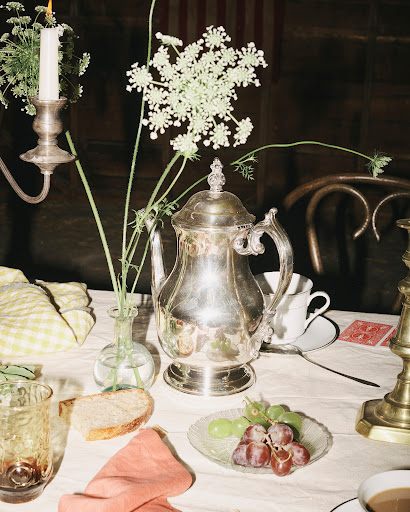 Still life of a silver teapot, antique glassware, bread, and grapes on a white tablecloth next to a candle, creating a warm and inviting atmosphere.