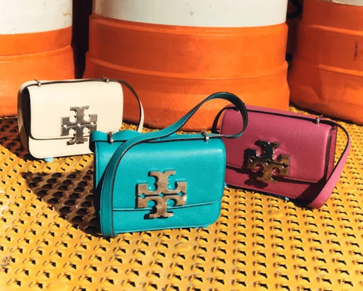Three Tory Burch handbags in white, teal, and pink, displayed on a yellow textured surface, with white and orange barrel-like objects in the background.