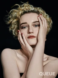 Close-up portrait of a woman with short curly blonde hair holding her hands to her face against a black background.