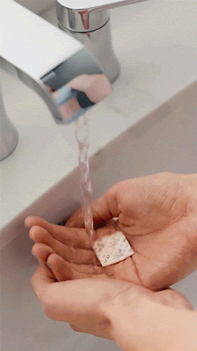 Person holding skincare patch under running water from a chrome faucet in a white porcelain bathroom sink.