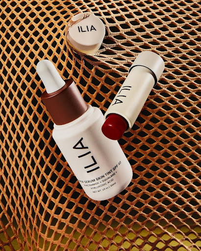 Three Ilia beauty products including a lip tint, serum skin tint, and a concealer, arranged on a gold mesh surface.