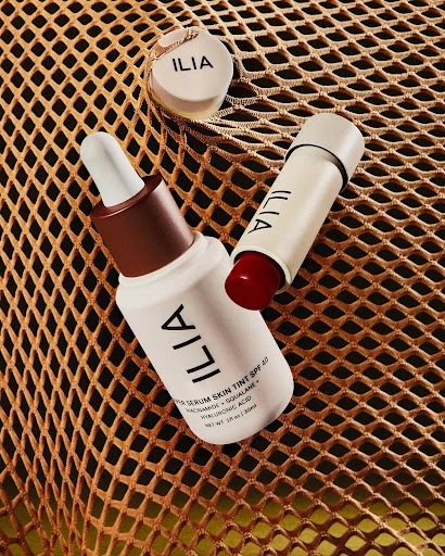 Three Ilia beauty products including a lip tint, serum skin tint, and a concealer, arranged on a gold mesh surface.