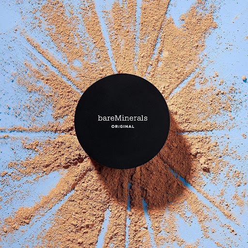 BareMinerals Original loose powder foundation with lid, tan powder surrounding the container on a light blue surface.