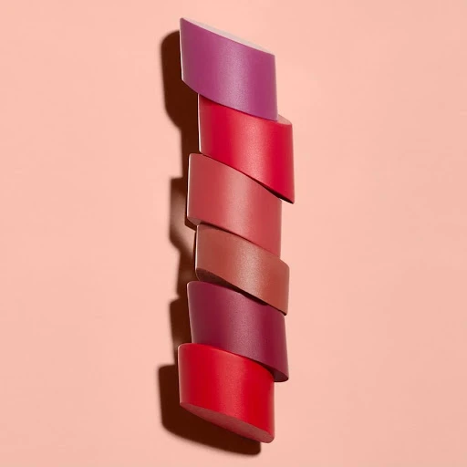 Tower of assorted lipstick samples in red, pink, and purple tones. Beauty advertising with product display on pink backdrop.