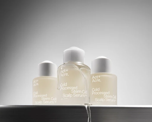 Three 'Act+Acre' bottles of Cold Processed Stem Cell Scalp Serum on a reflective surface with a gray gradient background. Beauty product.