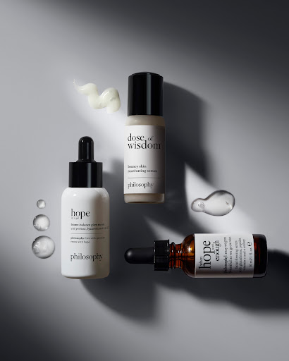 Three bottles of Philosophy skin serum arranged with clear serum drops and product smears on a light grey surface in studio lighting.