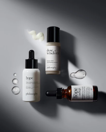 Three bottles of Philosophy skin serum arranged with clear serum drops and product smears on a light grey surface in studio lighting.