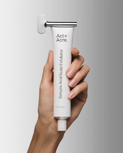 A hand holds a white tube of Act+Acre Salicylic Acid Scalp Exfoliator with a silver tube key against a gradient background.