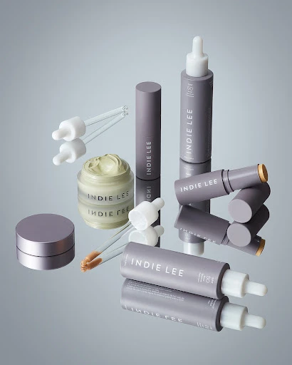 Various Indie Lee skincare products are displayed on a reflective surface against a gradient background. Jars, tubes, and dropper bottles.