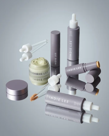 Various Indie Lee skincare products are displayed on a reflective surface against a gradient background. Jars, tubes, and dropper bottles.