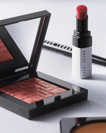 Close-up of Bobbi Brown makeup products, including a blush compact, lipstick, and eyeliner, on a white surface in soft, diffused lighting.