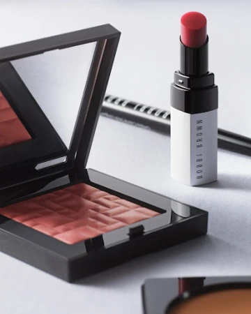 Close-up of Bobbi Brown makeup products, including a blush compact, lipstick, and eyeliner, on a white surface in soft, diffused lighting.