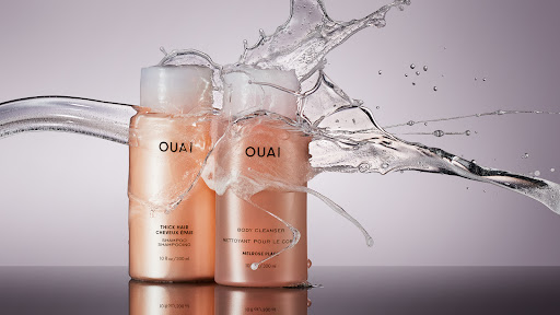 Two bottles of OUAI shampoo and body cleanser with water splashing against them, against a gradient background, beauty product photography.