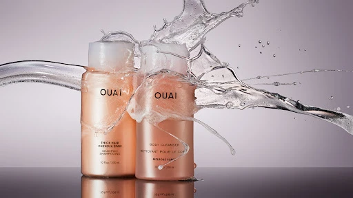 Two bottles of OUAI shampoo and body cleanser with water splashing against them, against a gradient background, beauty product photography.