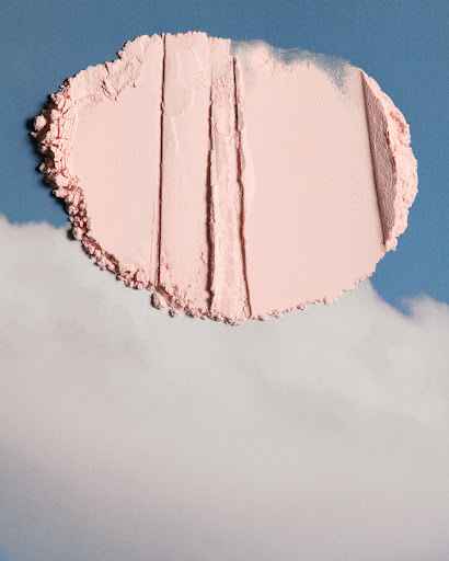 Pale pink powder makeup swatch on a sky-blue background with fluffy clouds. The powder has linear marks like a garden being raked.