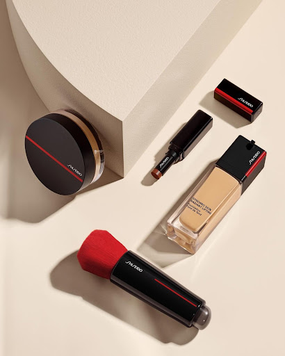 Overhead product shot of Shiseido makeup including foundation, powder, concealer, and brush on a light beige surface with soft shadows.