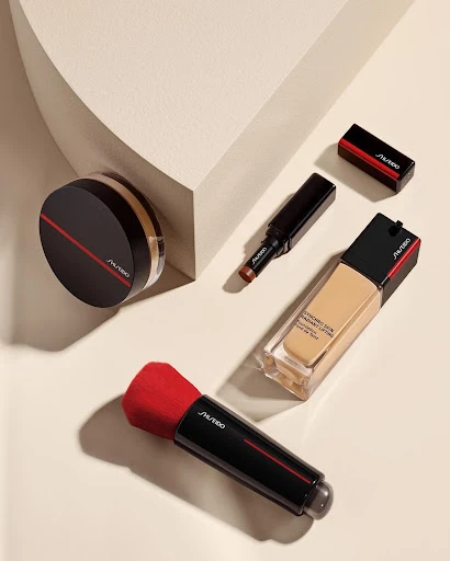 Overhead product shot of Shiseido makeup including foundation, powder, concealer, and brush on a light beige surface with soft shadows.