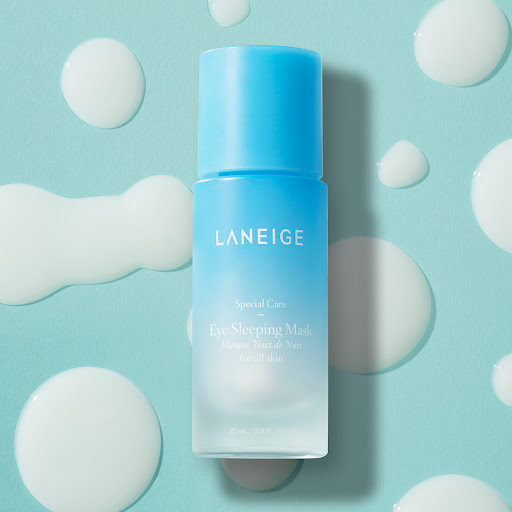 Close-up studio shot of a blue and white Laneige Eye Sleeping Mask container surrounded by simulated white liquid drops on blue backdrop.