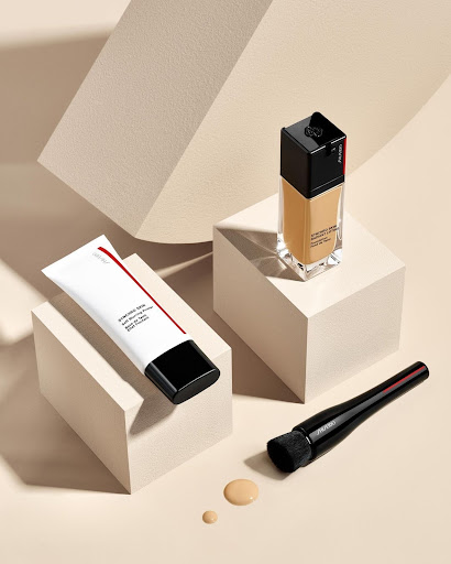 Still life product shot featuring a foundation bottle, primer tube, and makeup brush on geometric white blocks against a beige background.