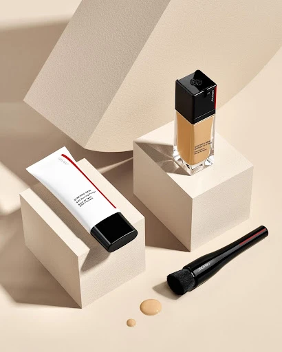 Still life product shot featuring a foundation bottle, primer tube, and makeup brush on geometric white blocks against a beige background.