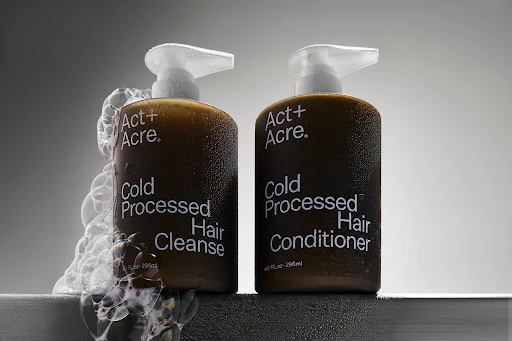 Two bottles of Act+Acre hair cleanser and conditioner, positioned side-by-side with pump dispensers, accented by cosmetic foam against a grey backdrop.