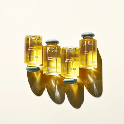 Four Origins Checks and Balances skincare bottles are positioned on a white surface, casting shadows. The bottles are filled with liquid.