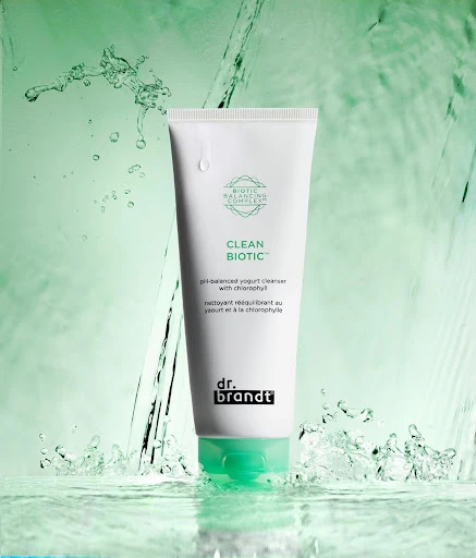A tube of Clean Biotic skincare product by Dr. Brandt on a reflective surface with water splashing around it against a light green backdrop.