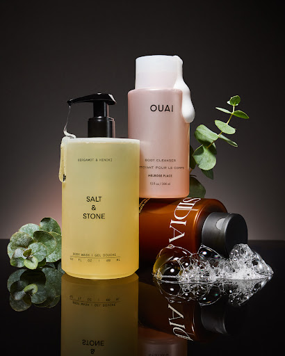 Still life shot of assorted body wash products from Salt & Stone and OUAI with sprigs of eucalyptus on a reflective black surface.