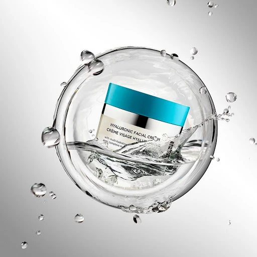 Facial cream jar with blue lid suspended in a splash of water against a gradient gray background, showcasing beauty and skincare.