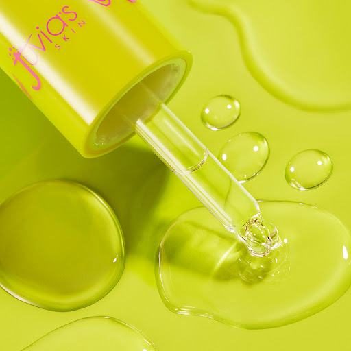 Close-up of a Juvia's Skin serum bottle and dropper with clear serum drops on a light green surface for beauty product promotion.