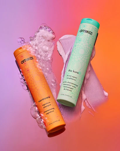 Two Amika brand shampoo and conditioner bottles are displayed against a pink and orange gradient background with scattered bubbles.