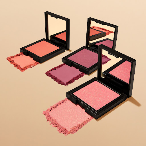 Three square compacts of blush are arranged on a tan surface, showcasing various shades of pink and coral makeup powder with mirrors.
