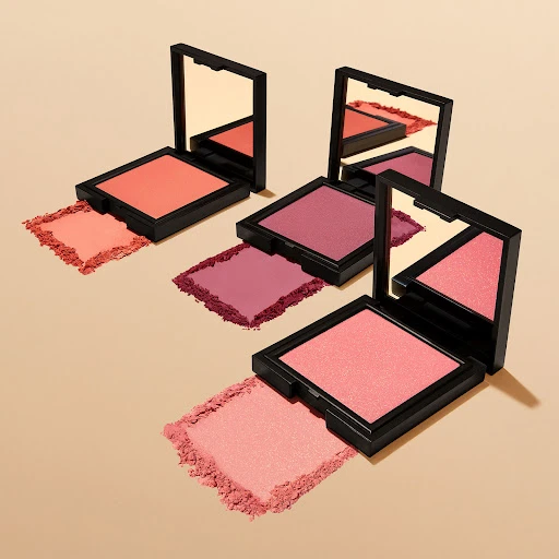 Three square compacts of blush are arranged on a tan surface, showcasing various shades of pink and coral makeup powder with mirrors.