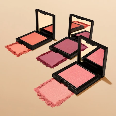 Three square compacts of blush are arranged on a tan surface, showcasing various shades of pink and coral makeup powder with mirrors.