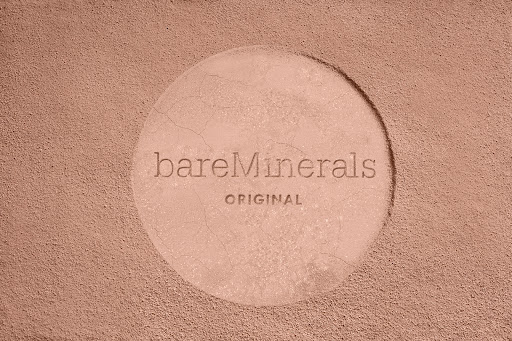 BareMinerals brand powder foundation with a circular imprint on textured tan background, product shot for beauty or fashion use.