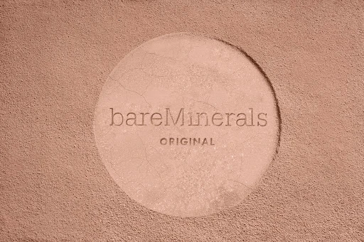 BareMinerals brand powder foundation with a circular imprint on textured tan background, product shot for beauty or fashion use.