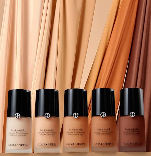Five bottles of Giorgio Armani Luminous Silk foundation in varying shades, lined up against a softly draped fabric backdrop in beige tones.