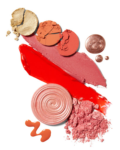 Overhead shot of assorted colorful makeup: eyeshadow, lipsticks, blush, and nail polish on a clean white surface in a beauty editorial.