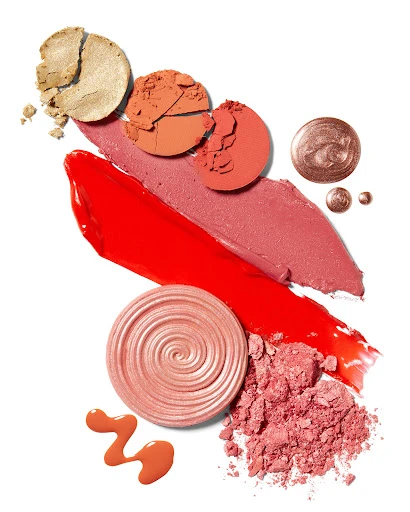 Overhead shot of assorted colorful makeup: eyeshadow, lipsticks, blush, and nail polish on a clean white surface in a beauty editorial.