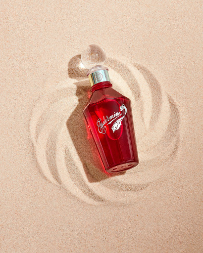 Red glass bottle of liquid fragrance on sand with circular pattern, shot in a photo studio with soft lighting.