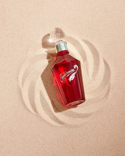 Red glass bottle of liquid fragrance on sand with circular pattern, shot in a photo studio with soft lighting.
