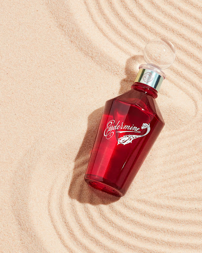Red glass bottle with clear spherical stopper set on tan sand with concentric wave patterns and side lighting for visual appeal.